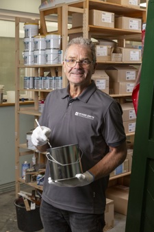 Wyger Smits poseert in een grijze polo met de naam van zijn bedrijf erop. Hij heeft kort grijs haar en draagt een bril. Hij lacht en staat tussen de schappen met blikken verf.