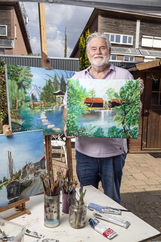 Hans Boetzkes staat op de foto met zijn olieverfschilderijen. Hij kijkt vriendelijk, heeft grijs haar en een grijze baard. Op het schilderij in zijn handen staat een huis aan een meer.