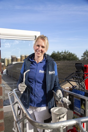 Op de foto staat Marloes. Marloes is een blonde vrouw met een blauwe trui aan waarop "Baars schilderwerken" staat. Ze heeft haar haren in een knot en lacht breed. In haar hand houdt ze een kwast en ze poseert op een hoogwerker. De lucht achter Marloes is blauw.