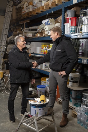 Wyger Smits poseert met zijn opvolger Jasper van Veen. Ze staan samen tussen de schildersspullen. Wyger schudt Jasper de hand. Ze lachen allebei. Jasper is een lange man met blond haar. Hij draagt een zwarte jas met de naam van het bedrijf erop.