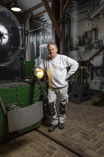 Nol Grasman poseert naast een zwart/groene locomotief. Hij is een man met grijs haar en een witte trui aan. Hij draagt ook een schildersbroek.