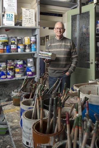 Jan van Wijnen poseert tussen kasten met potten met verf. In zijn hand houdt hij een stapel boeken. Jan draagt een bril, heeft grijs kort haar en een groen gestreepte trui aan. Hij lacht.